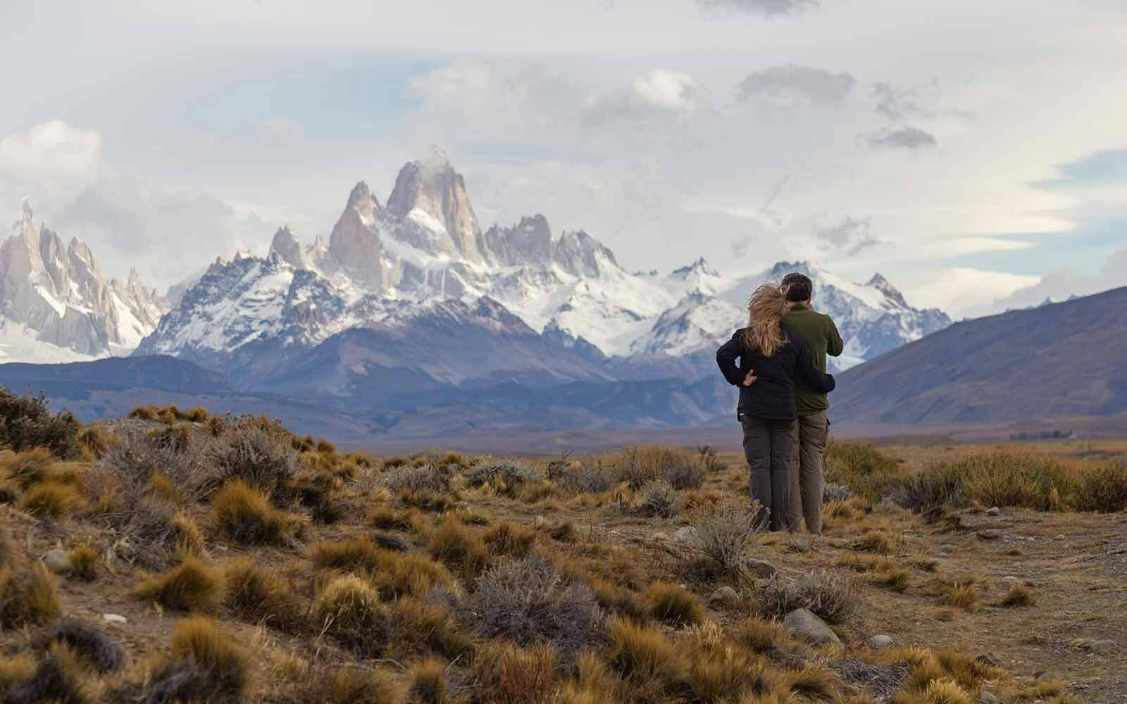 Two people standing in a field with mountains in the background.