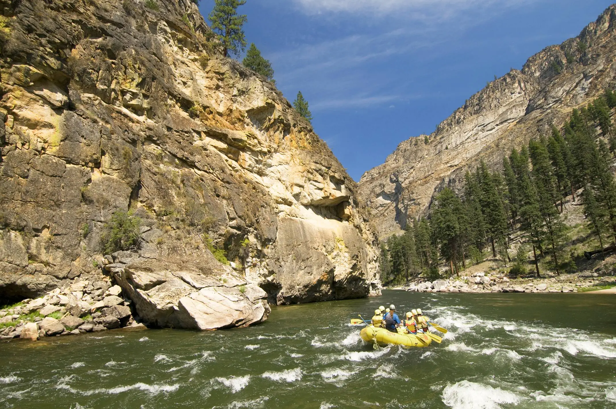 A group of people in a yellow inflatable raft paddle down a river surrounded by rocky cliffs and pine trees under a blue sky, enjoying one of the area's thrilling River Rafting & Kayaking Tours.