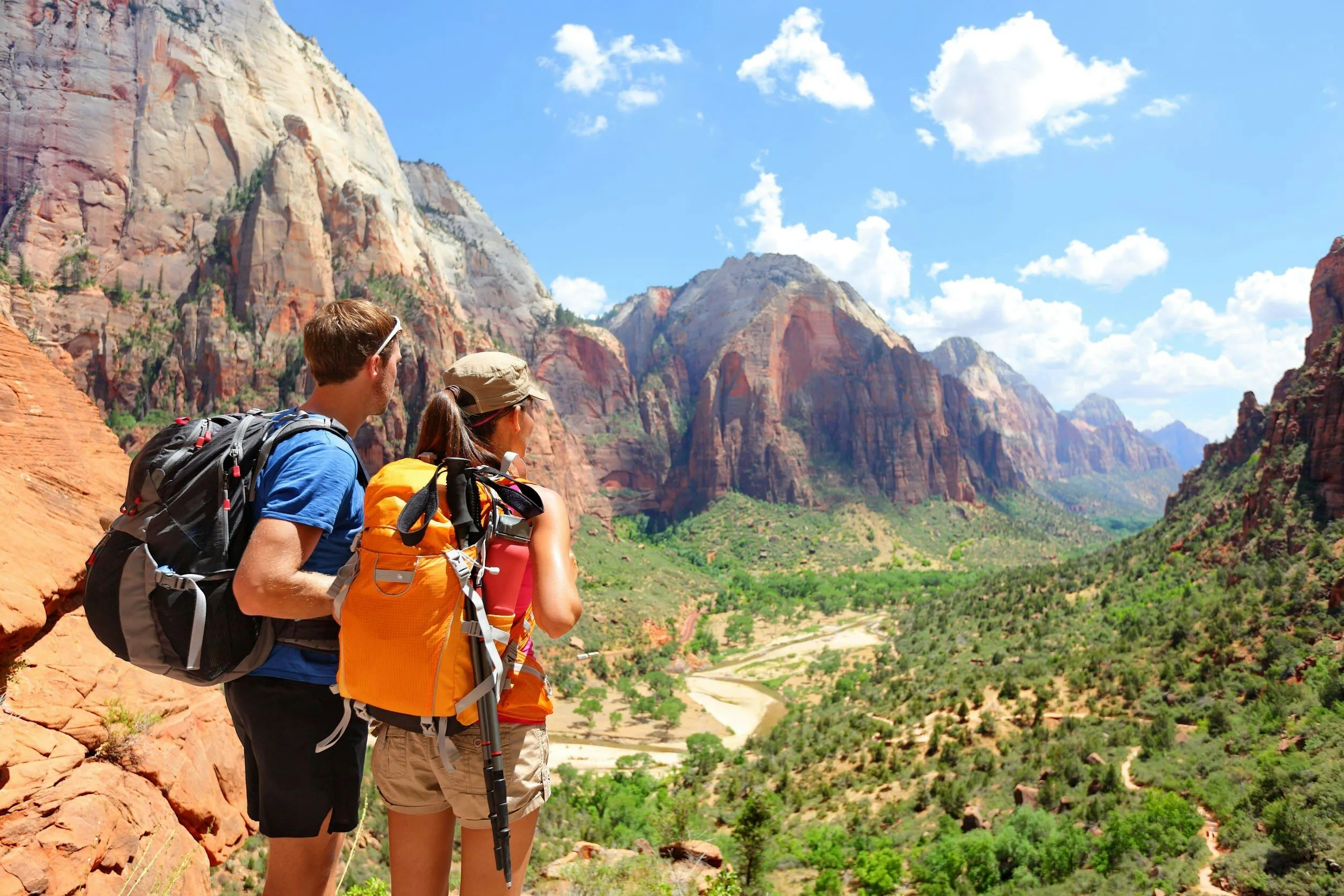 Two hikers with backpacks stand on a rocky ledge overlooking a scenic canyon, reflecting on recent REI cancelled trips as they admire the rugged cliffs, green valley, and winding river beneath a partly cloudy sky.