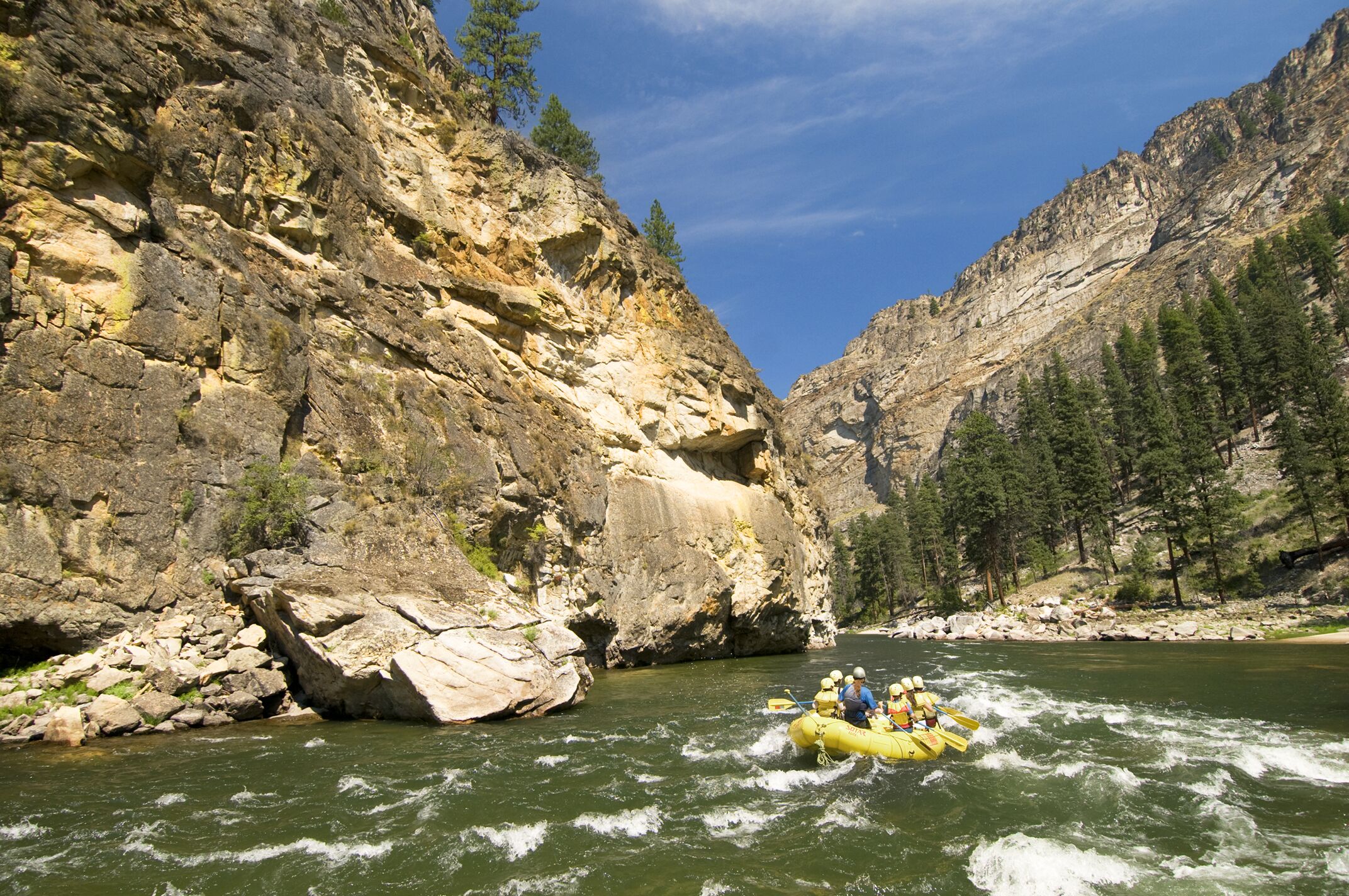 A group of people in a yellow inflatable raft paddle down a river surrounded by rocky cliffs and pine trees under a blue sky, enjoying one of the area's thrilling River Rafting & Kayaking Tours.