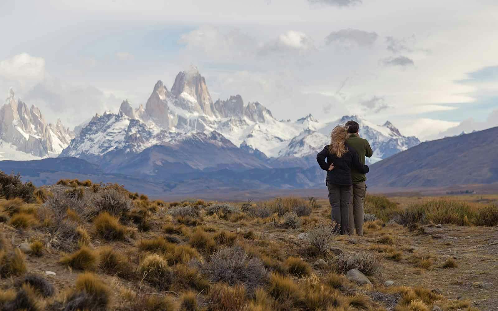 Two people standing in a field with mountains in the background.