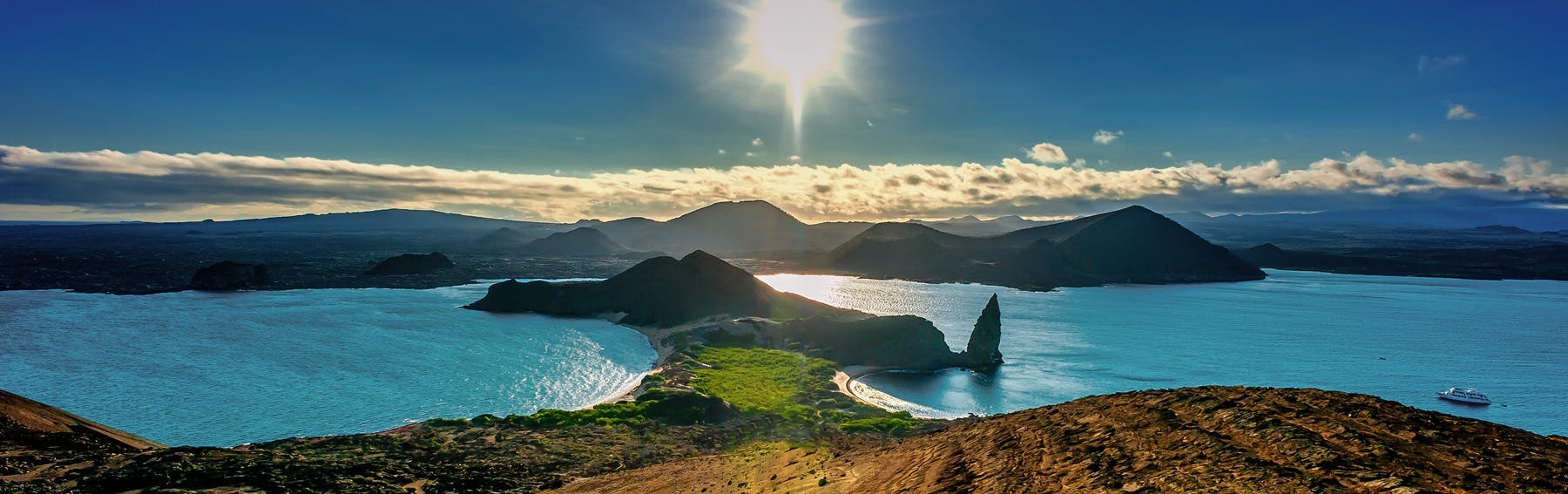 A panoramic view of a coastal landscape at sunset, featuring blue water, a small peninsula, rocky hills, and a distant boat on the right—perfect for winter adventures along the serene shoreline.