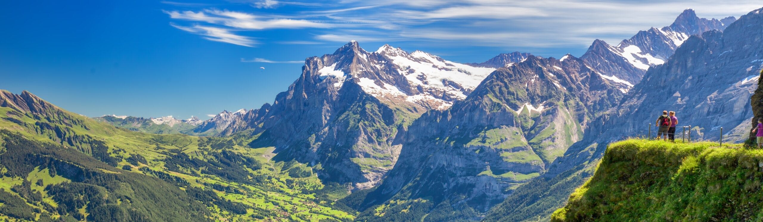 Snow-capped mountains and green valleys under a blue sky, with a few hikers standing on a grassy ledge overlooking the landscape—an unforgettable moment from hiking in Europe.